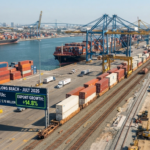 A realistic, wide-angle aerial view of the Port of Long Beach during daytime, showing container ships docked at terminals, stacked shipping containers, and a freight train moving along on-dock rail tracks alongside digital display boards showing port volume statistics.